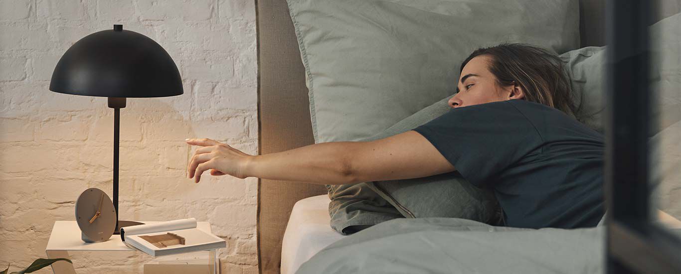 Femme au lit s'étire vers la lampe de chevet. 